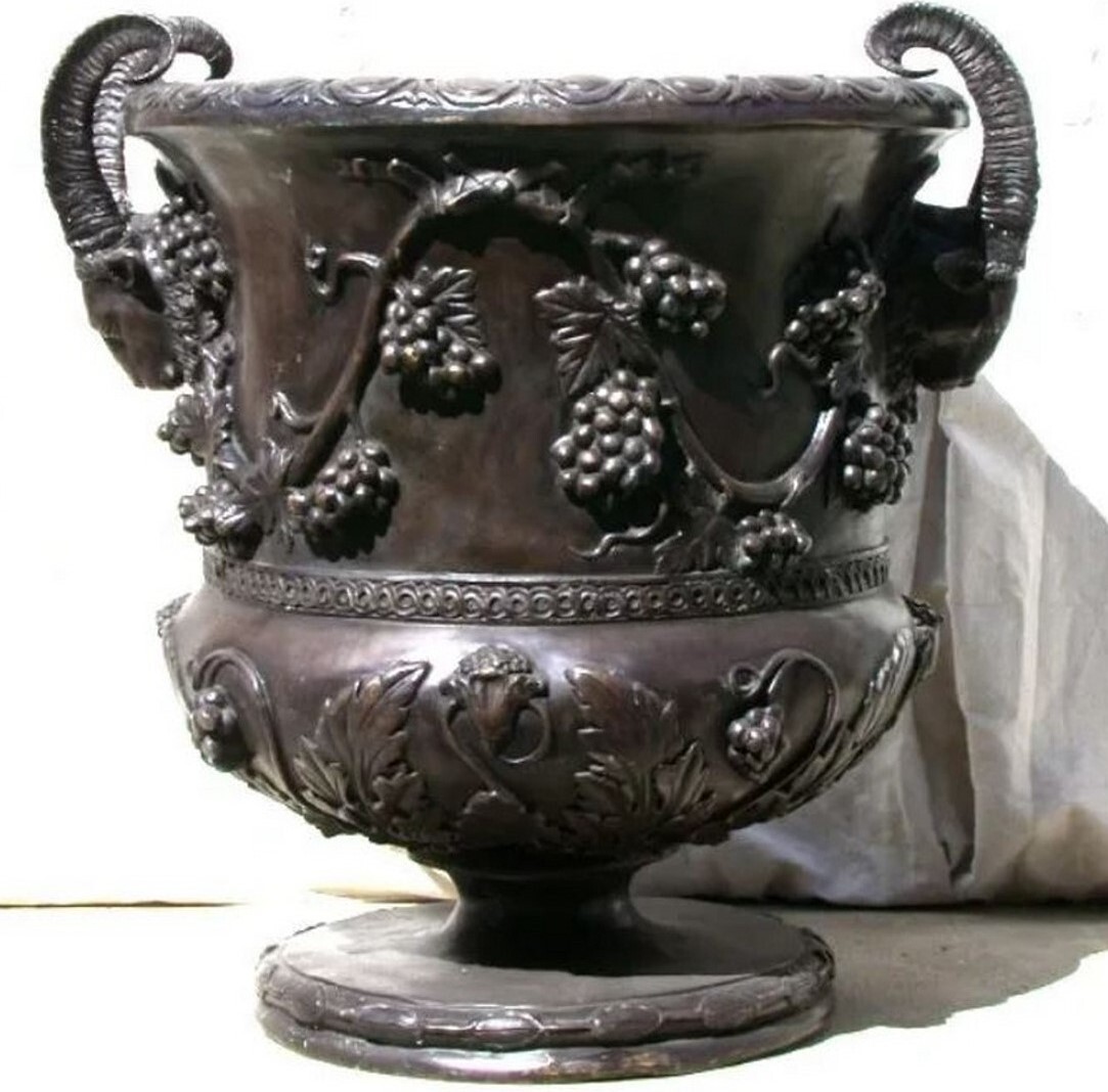 This classical Roman-style bronze planter urn features a pair of rams head handles and decorative grapevine accents.